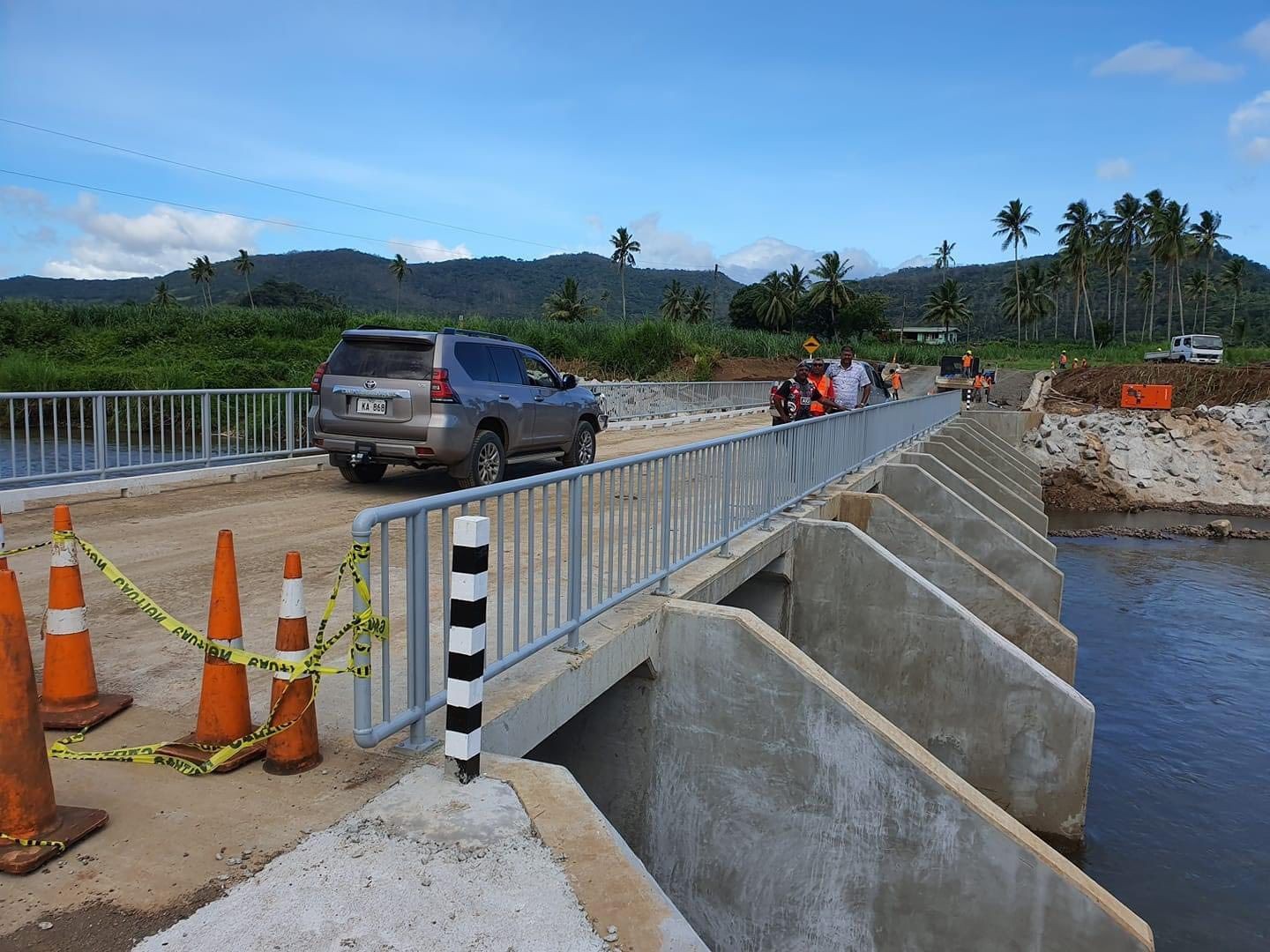 Vehicles and people crossing a concrete bridge surrounded by cones in a tropical landscape.