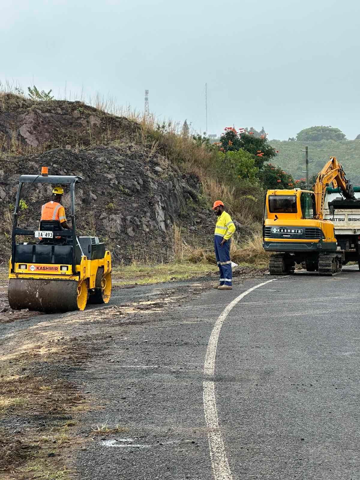 Construction workers using machinery to pave a road near a rocky hill and vegetation on a cloudy day.