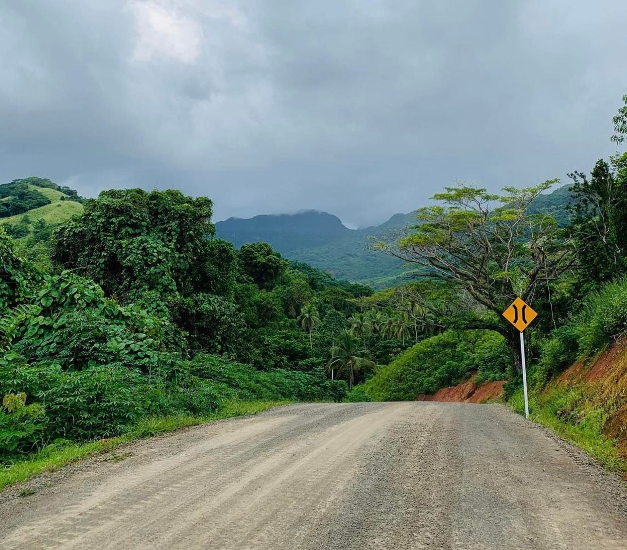 Dirt road leading through lush green hills with overcast sky and a warning road sign.